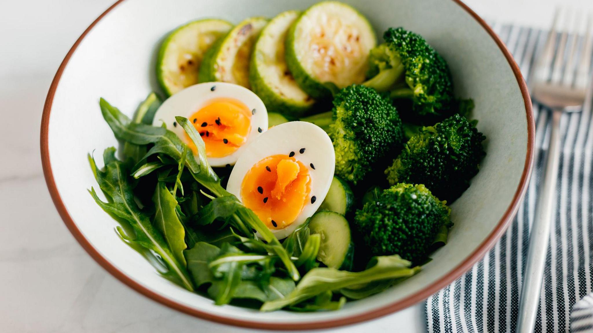 A bowl with a hard boiled egg sliced in half, with salad leaves, broccoli and cucumber slices.