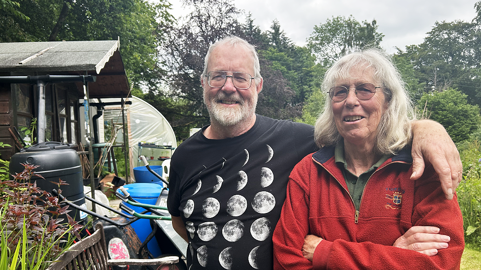 Ken O'Brien and Susan Lancaster are a white man and woman in their 70s. Ken has white hair and a white beard and wears wire-framed rectangular glasses and a black t-shirt with different phases of the moon photographs across it. Susan has shoulder length white hair and a fringe and wears slightly tinted glasses. She wears a green polo shirt with a red three-quarter zip fleece top with a crest to the left side of the chest. She has her arms crossed and he has one arm over her shoulders. They are both smiling at the camera. Behind them are trees, the edges of an allotment growing area, a shed with multiple plastic composters and containers and tools outside, a wooden bench and a polytunnel in the background. The photo is framed by trees.