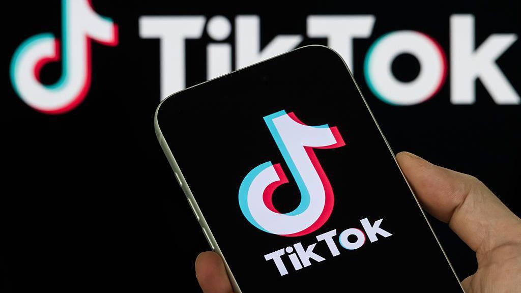 A smartphone held in someone's hand displays the TikTok logo, with a backdrop of the company's logo behind it