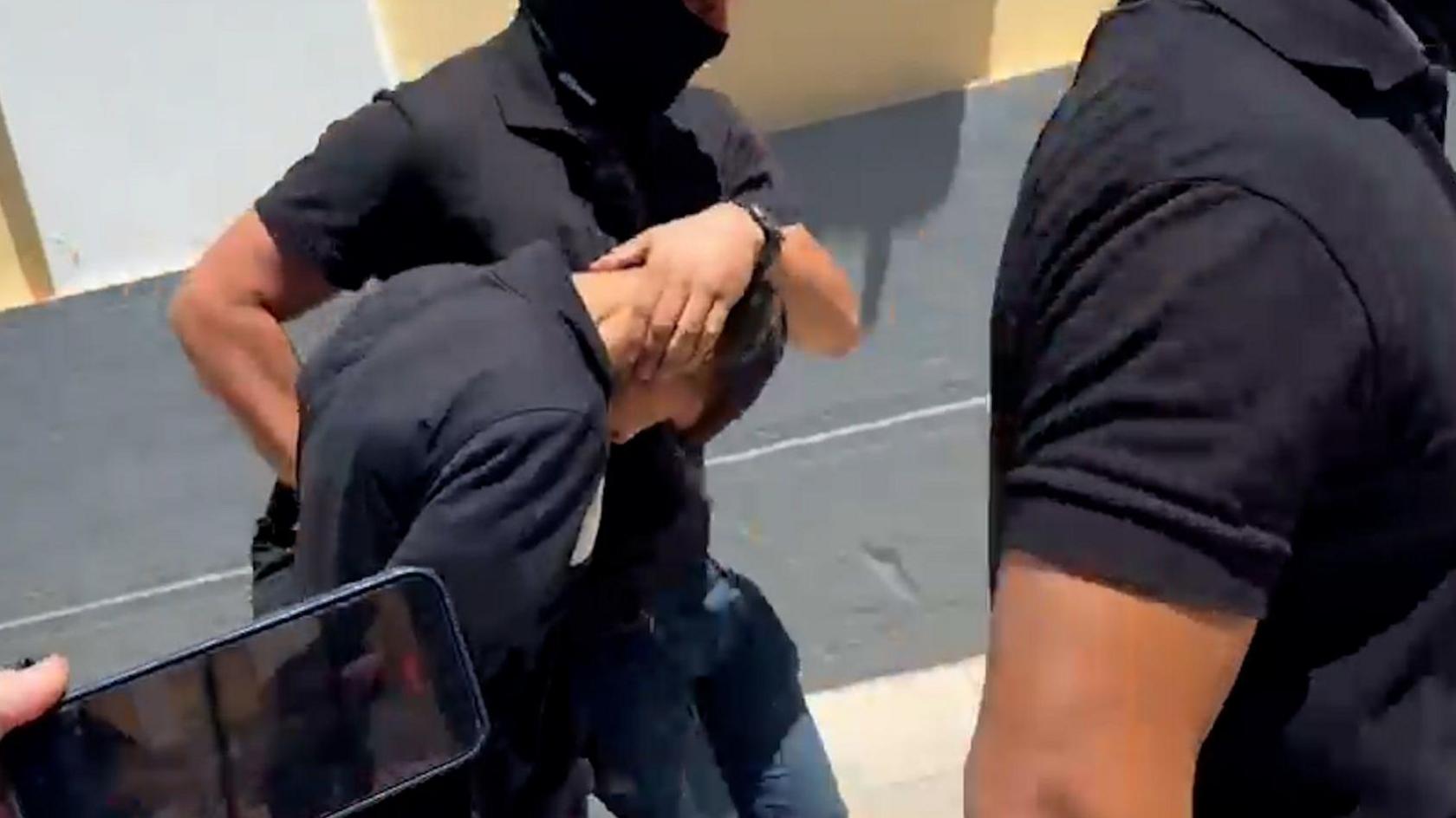 A man with his head down flanked by two police officers with their faces covered.