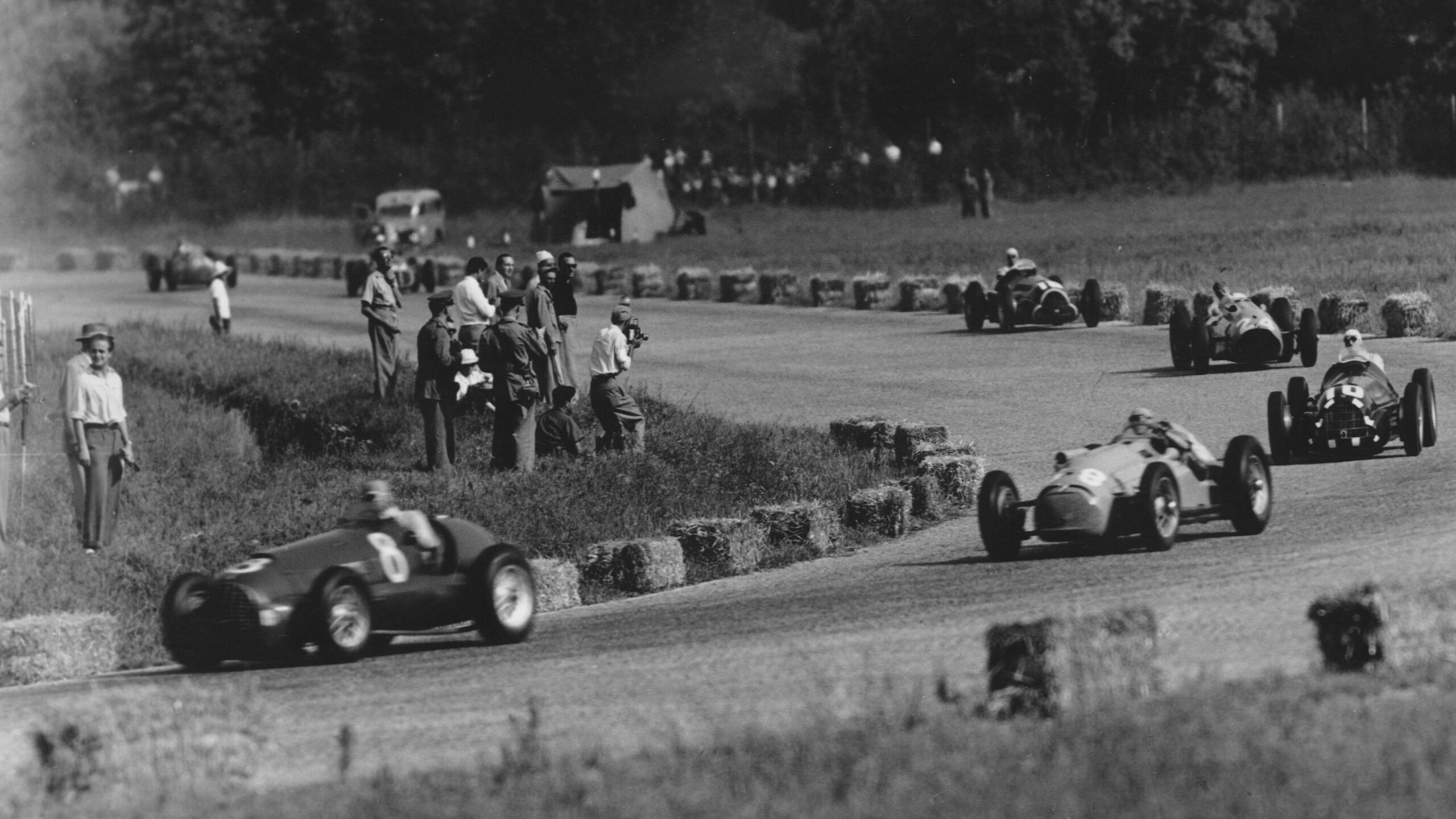 Juan Manuel Fangio leads the 1950 Italian Grand Prix. There are many people and photographers gathered on the inside of the corner