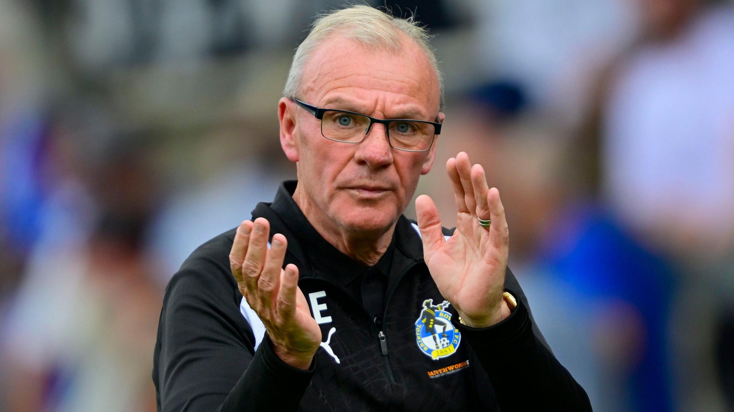 Steve Evans, wearing a black Bristol Rovers tracksuit top, claps his hands after the EFL Cup loss to Peterborough United