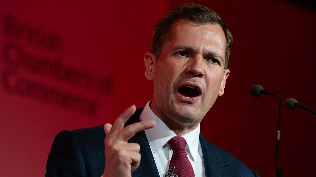 Robert Jenrick makes a hand expression as he speaks at a podium in front of a red-lit background.