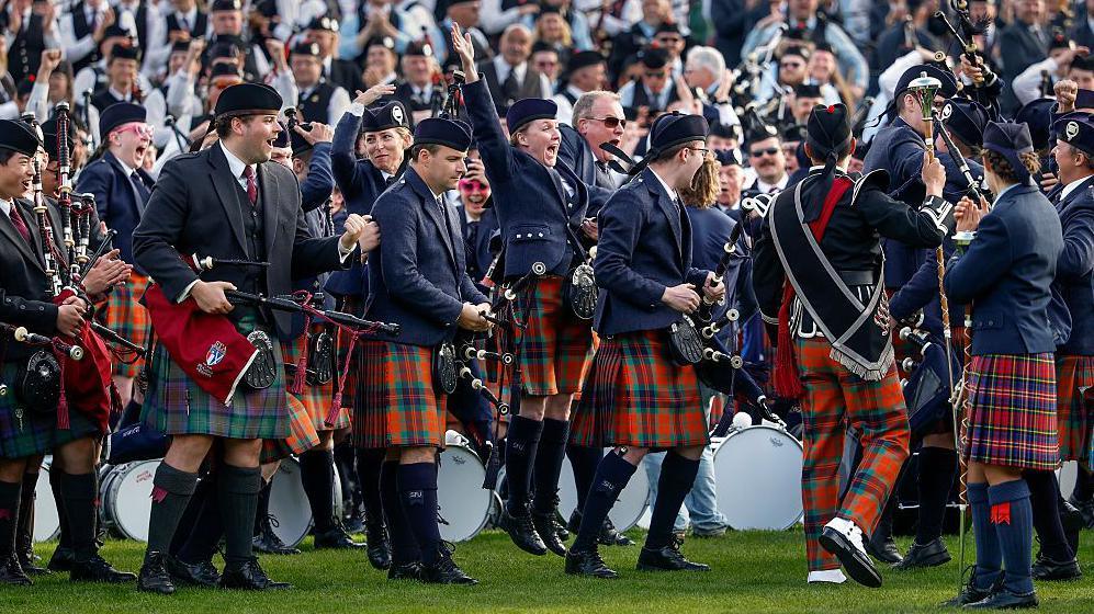 Members of the Simon Fraser University pipe band celebrating their win. Some are shouting with joy and raising their hands in the air. They are wearing traditional piper outfits with hats, A woman in the middle is jumping in the air