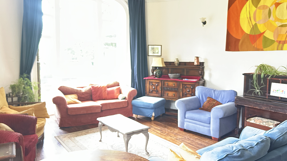 A large sunny sitting room with two sofas and two armchairs,a footstool, a piano, a dark wooden sideboard, a number of coffee tables and a large printed orange, yellow, green and brown piece of fabric on the wall. there are a couple of plants on top of the piano and side tables. There is a large arched floor-length window at the back of the photo framed by long blue velvet curtains.