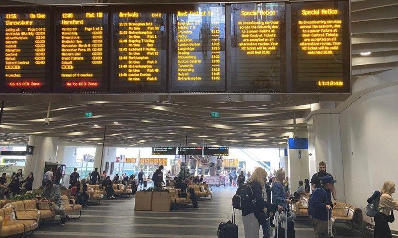 There is a series of digital information screens on rail services hanging from the ceiling at a station, with passengers waiting on the concourse, some on seats provided.