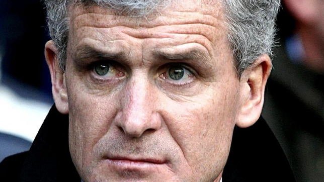 Image shows Mark Hughes, who has grey hair and is wearing a black overcoat, sitting in the stands of a football stadium