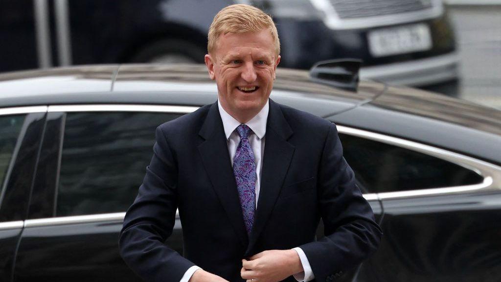 A man in a suit with a purple blue tie smiles in front of a black car.