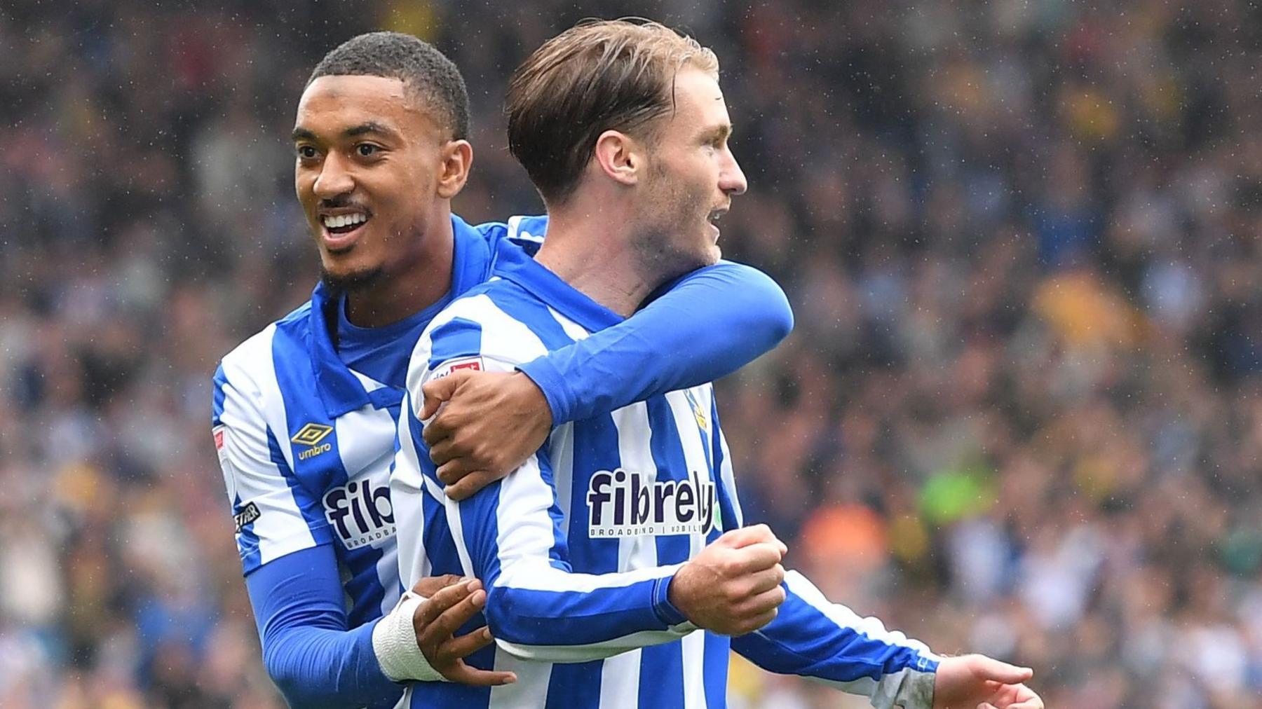 Slattery pumps right fist and looks over his left shoulder as a fellow player throws his arm araound him in celebration, wearing Wednesday blue and white striped home kit