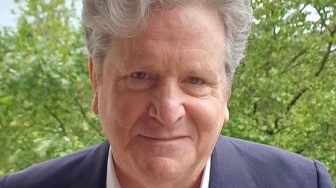 John Stevens smiles for the camera. He has thick grey hair and is wearing a blue jacket and open necked white shirt.
