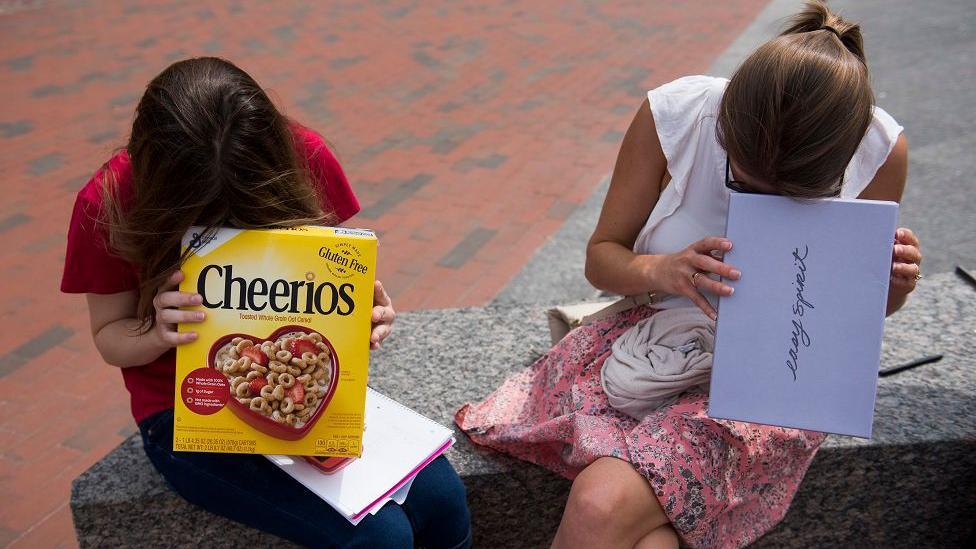 Two girls look into cardboard boxes, one a cereal box and another a shoe box, while sitting on a bench outside.