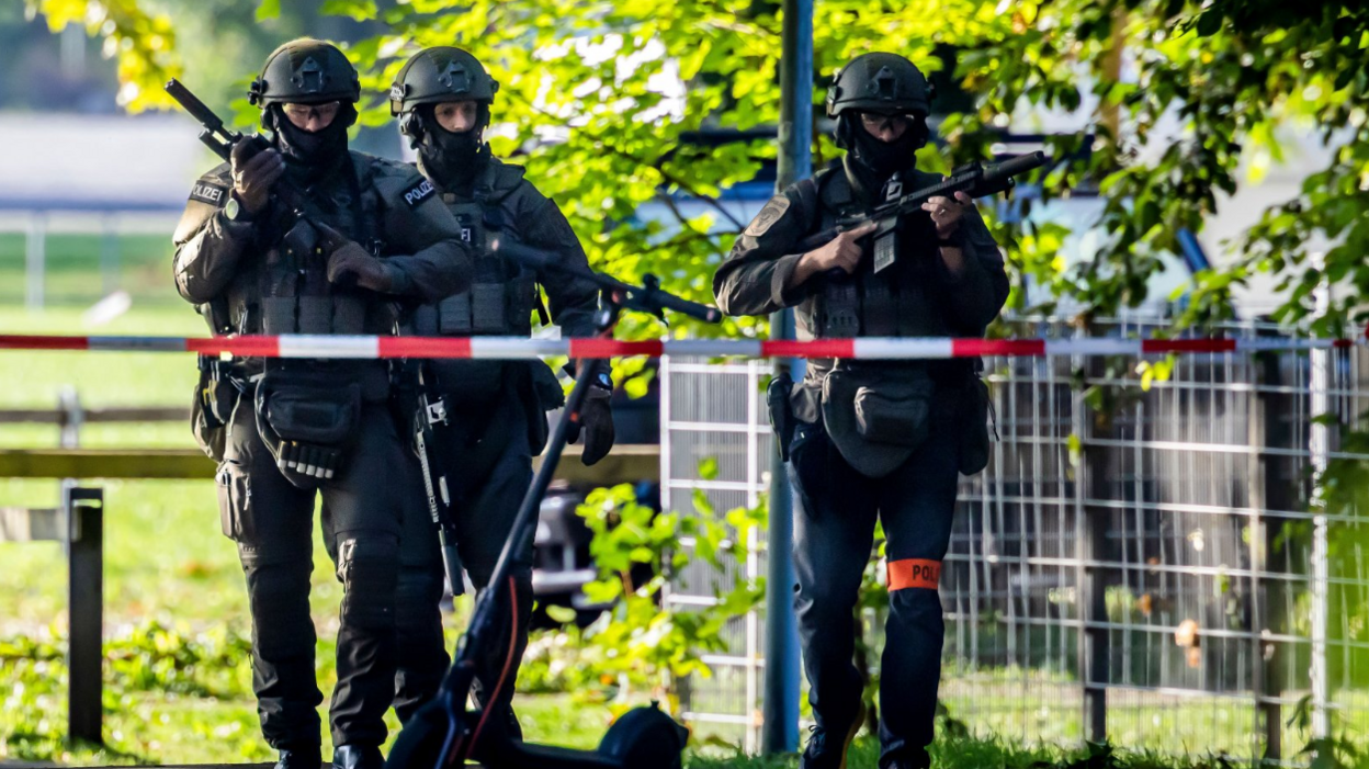 Three police officers in riot gear (helmet, bulletproof vest, knee and elbow protection) carrying guns patrol on the street, a police cordon is visible in the foreground