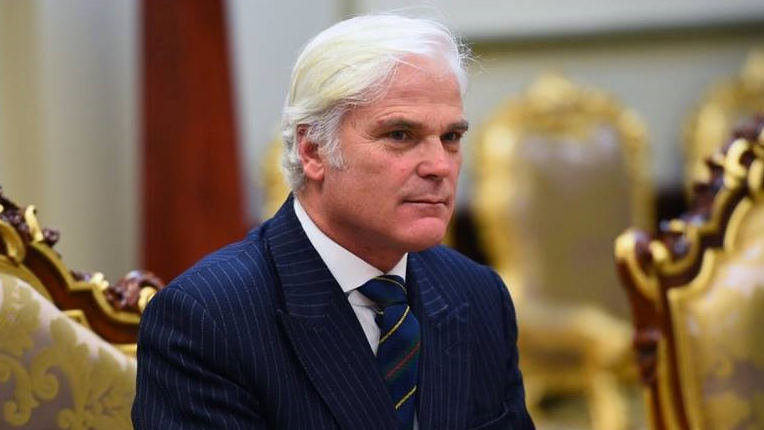 Headshot of Sir Desmond sitting in a room with gold chairs. He has medium length white hair and wears a navy pinstripe suit with a white shirt and a navy and gold tie.