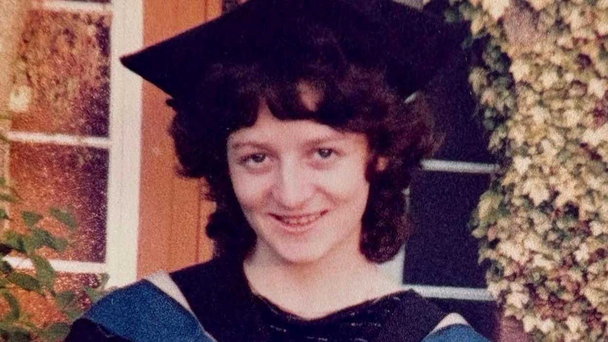 Linda smiles at the camera wearing a graduation cap and gown. She has short, dark curly hair and is outside a house.