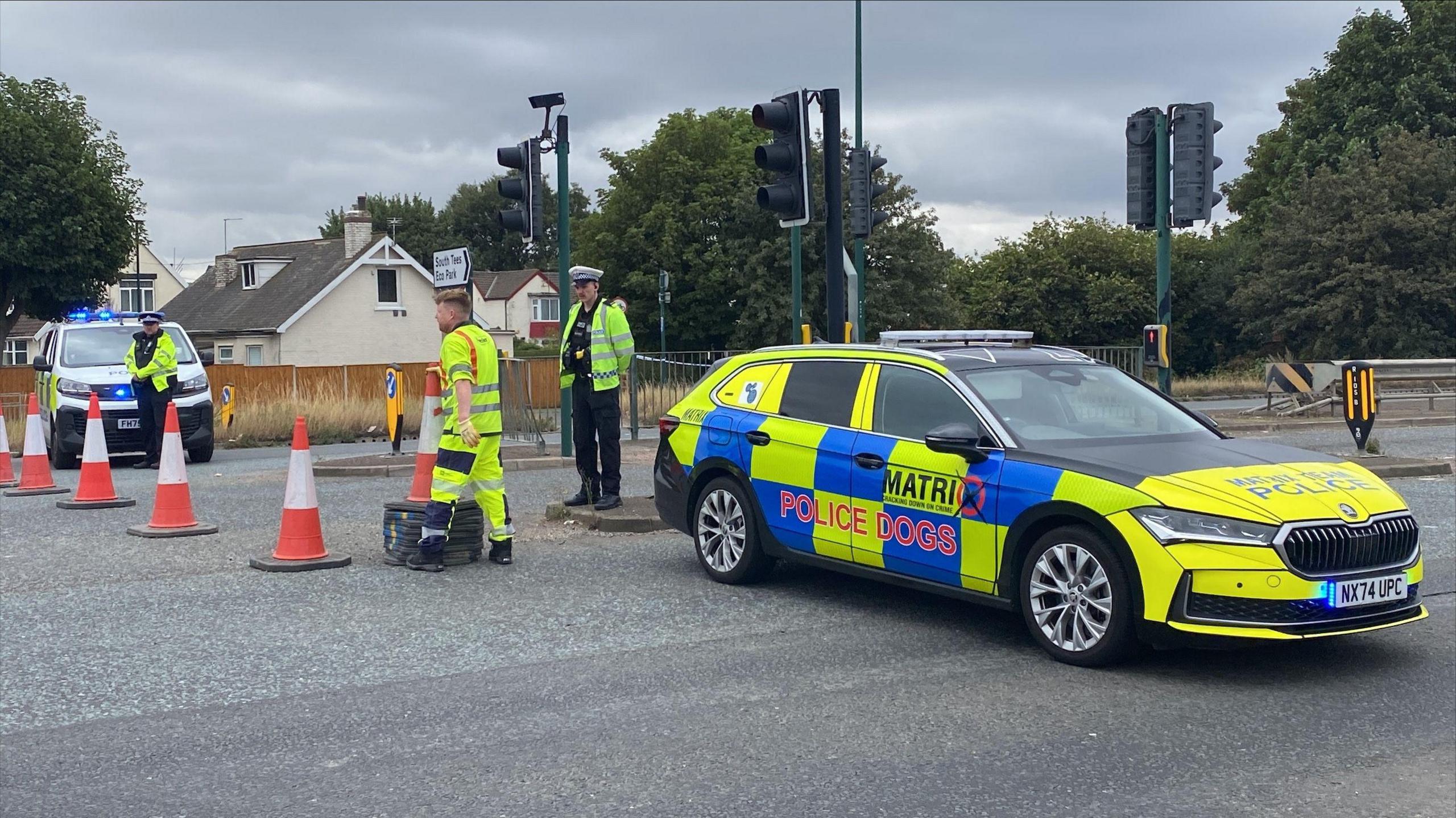Where the road has been closed, a marked police car with "Police Dogs" written on it is parked across the lane, blocking it. A police van with flashing blue lights is parked nearby. A man wearing high-visibility overalls is laying cones out in a straight line across the road. Two more officers stand nearby in uniform, one with their hands behind their back.