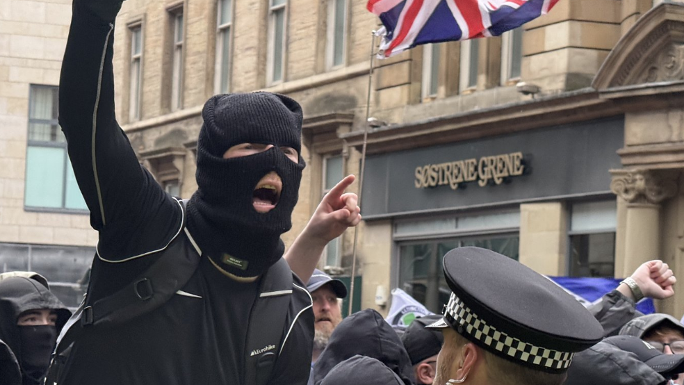 A masked individual in dark clothing raises a clenched fist while standing close to a police officer in a high-visibility jacket and checkered hat, with a crowd gathered behind and a Union Jack flag visible above on a city street lined with stone buildings and shopfront signage reading “Sostrene Grene.”