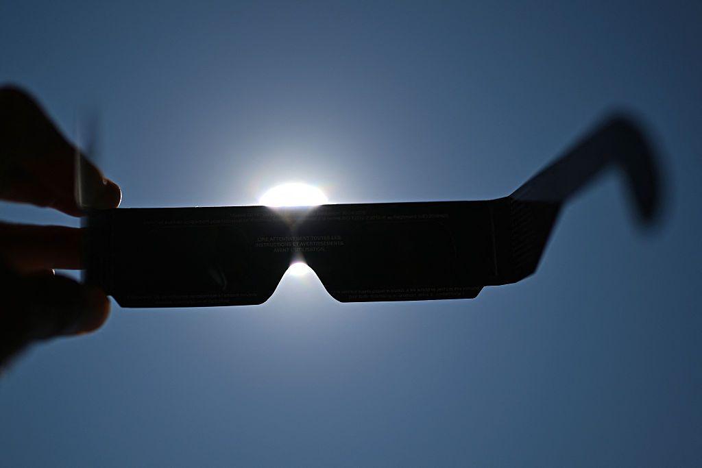 Hand holding up eclipse-viewing glasses to a blue sky