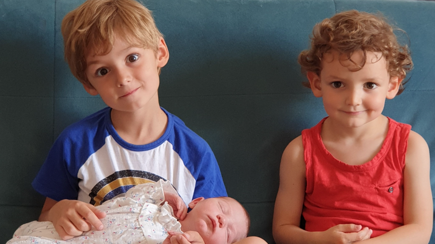 Two young children aged approximately three to five years old are sitting on a sofa with a newborn baby lying across the older child's lap. He is a boy who is staring at the camera with a smile (no teeth showing), his head tilted to the side. The younger child next to him is a girl. There is a close family likeness between the two, especially in their brown eyes.