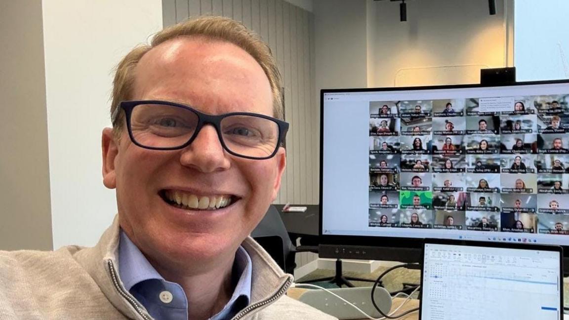 Man wearing glasses smiling at camera with computer screen behind him