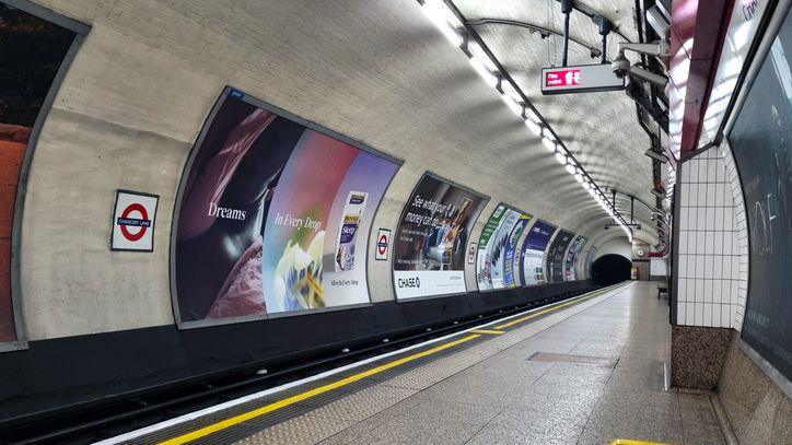 An empty platform at Chancery Lane Underground station in London, looking down the curved track tunnel. Large advertising billboards line the tiled wall opposite the platform, illuminated by overhead strip lighting. A dark tunnel entrance is visible in the background, with a London Underground roundel sign mounted on the left wall.