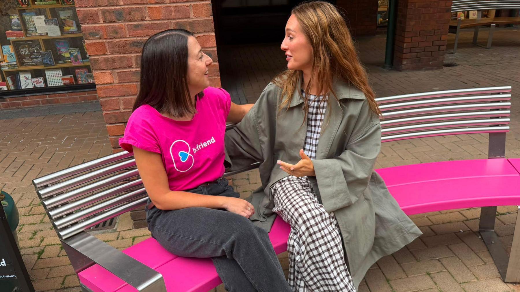 Two women are sitting on a curved bench with a metal backrest and a bright pink seat. They are turned towards each other and are smiling. The woman sitting on the left has shoulder length dark hair. She is wearing a bright pink t-shirt with the word b:friend on it and black jeans. The other woman has longer light brown hair and is wearing a grey mac over a black and white ankle length dress. They look like they are in the middle of a conversation.