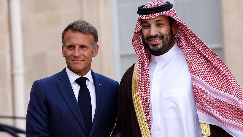 A smiling Macron, wearing a navy suit and black tie, poses next to bin Salman, who is wearing traditional Saudi attire