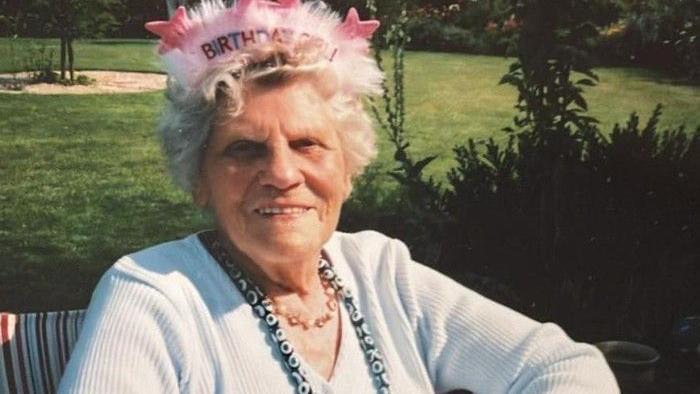 An old woman in a cardigan and necklace sitting outside wearing a hat that says Birthday Girl! on it.