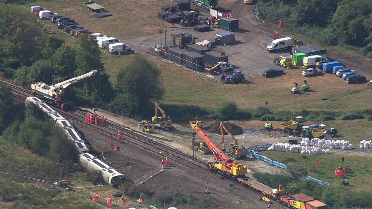 An aerial view of the train derailment site near Lewes shows several carriages spread across and beside the railway line. Recovery crews in orange high-visibility clothing are working around large cranes, excavators and engineering vehicles. Temporary compounds, parked vans, equipment and stacks of white bags are visible beside the track.