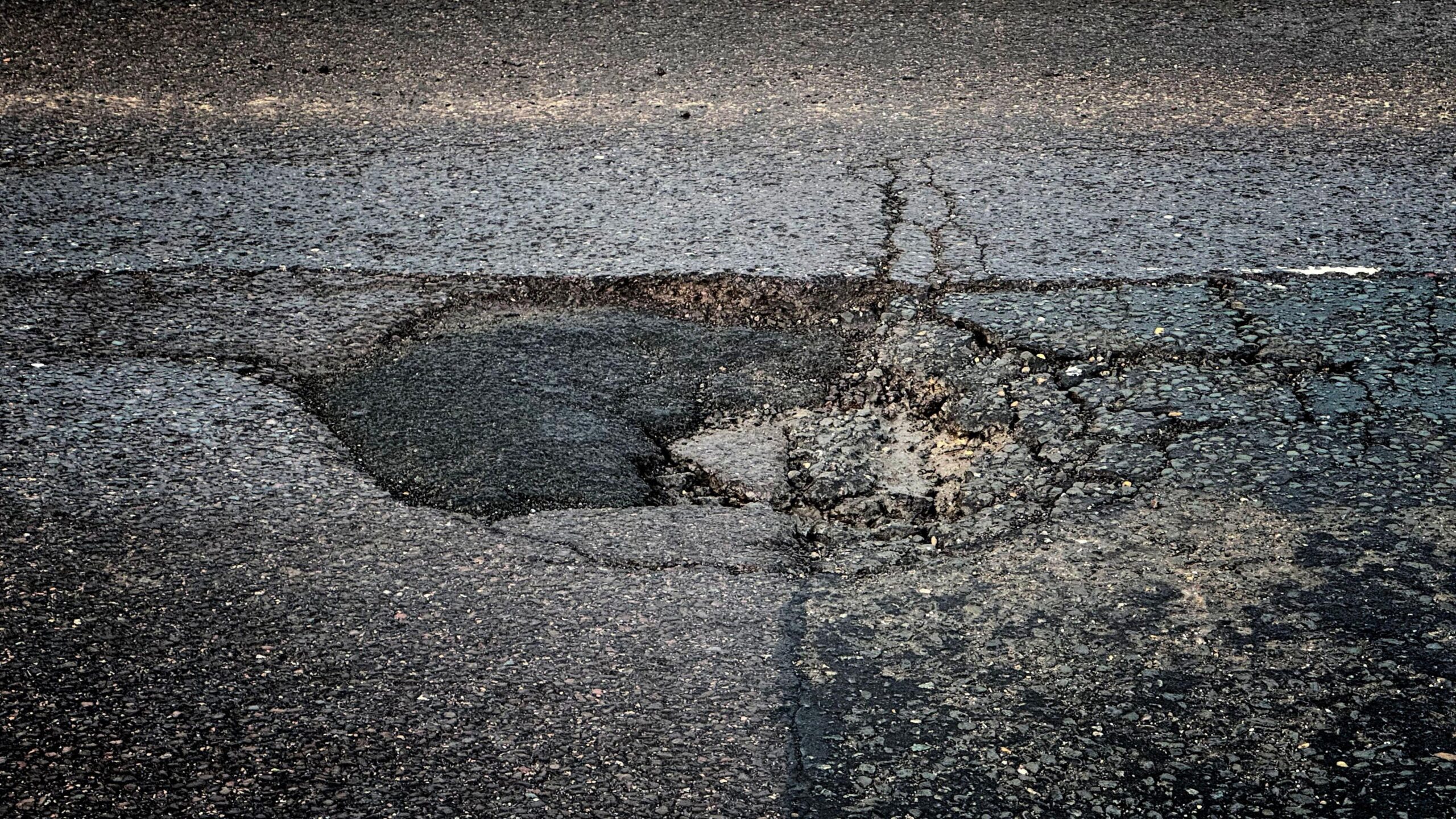 Close-up image of a pothole. It is roughly circular with a number of cracks, and part of it looks to have been filled with asphalt, which is darker in colour than the surrounding road surface.