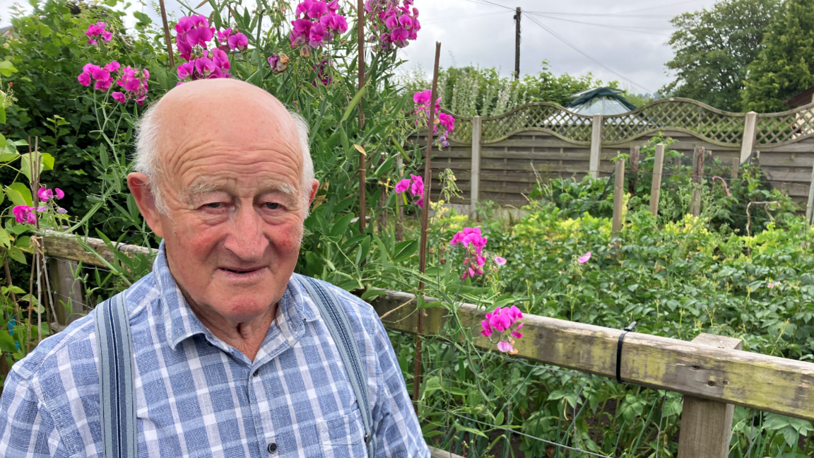 Meredydd, an elderly man in a blue check shirt looking into the camera. He's smiling and is standing in front of a garden with pink flowers.