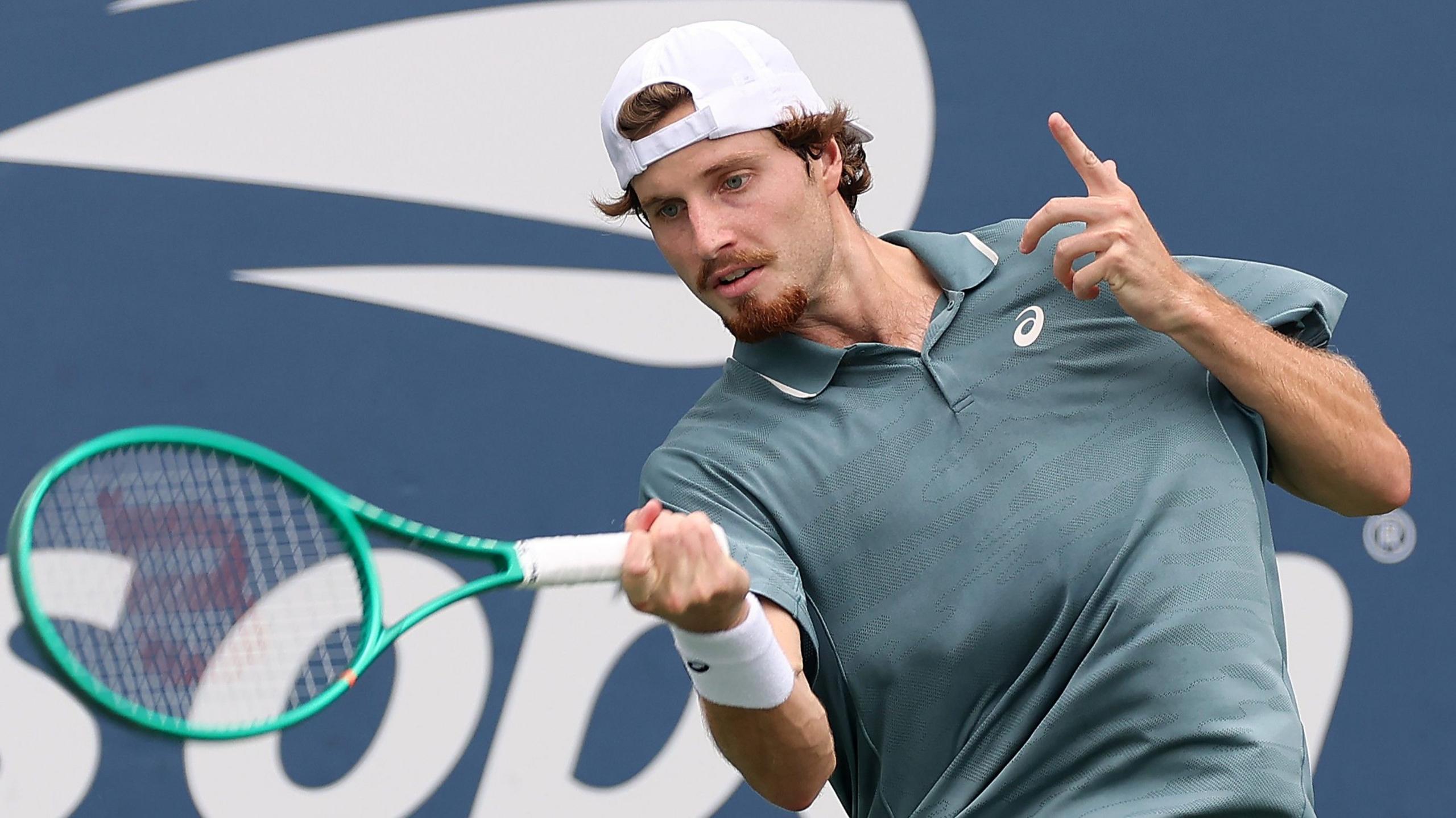 Toby Samuel, wearing a grey shirt and white backwards baseball cap, hits the ball with a green racquet