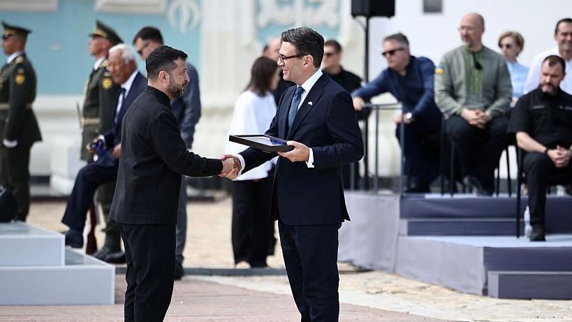 Zelensky and Burnham shake hands and smile as the latter holds a frame