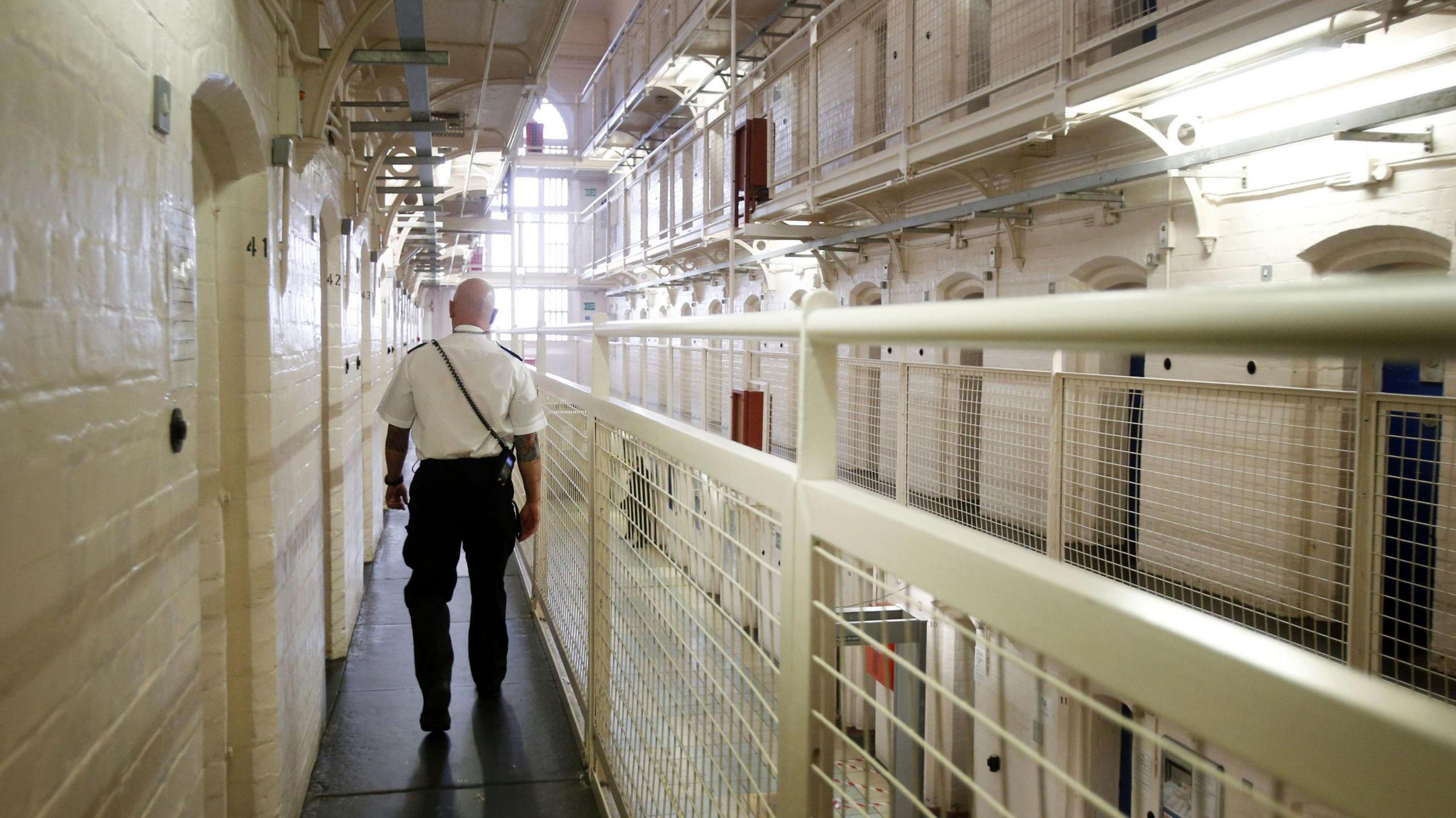 Prison Guard stands in a prison with his back to the camera he is wearing a white shirt and black trousers