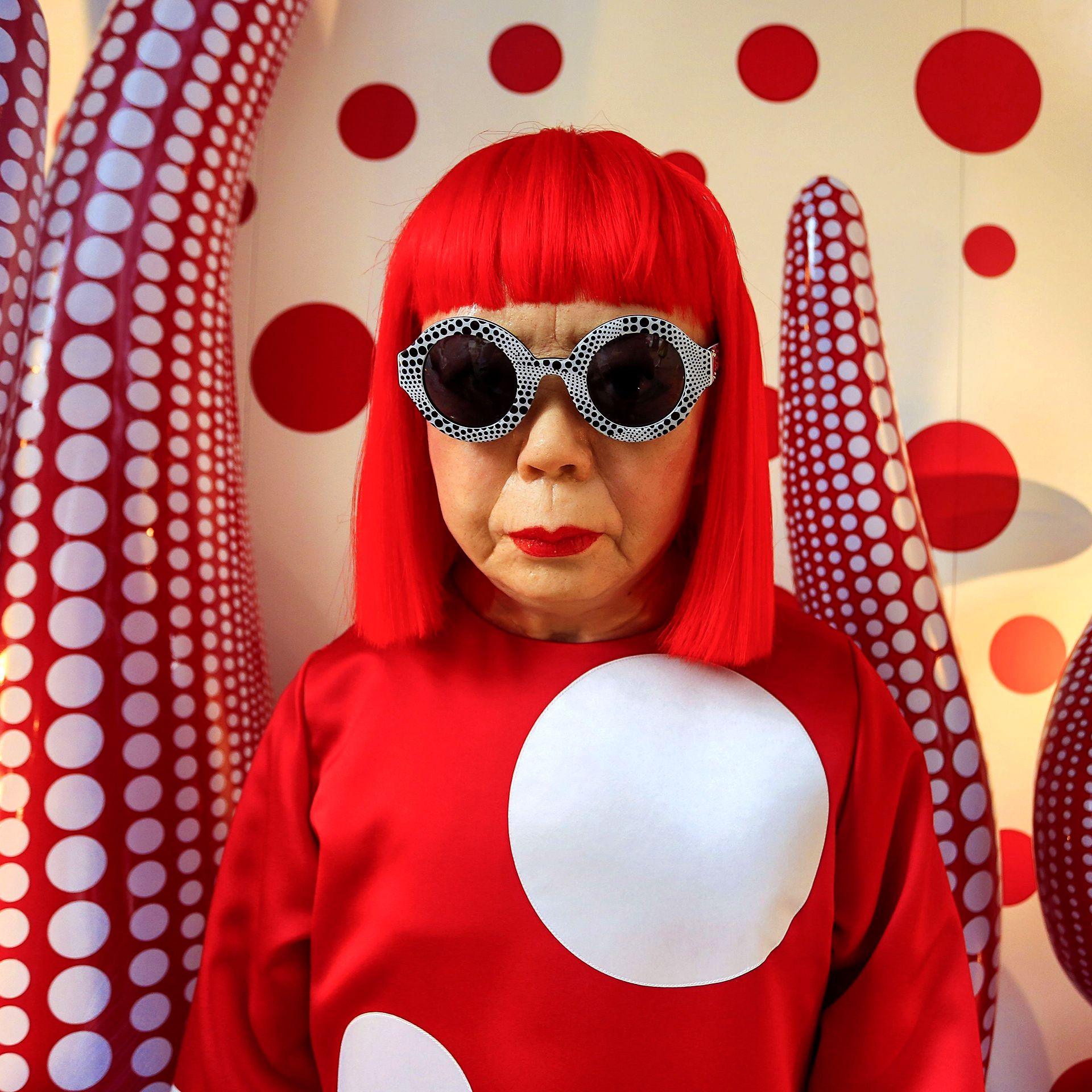 Yayoi Kusama looks at the camera wearing round glasses and a red top with white polka against a backdrop of polka dot tentacles for her exhibition in Hong Kong in 2012.