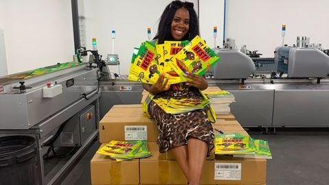 Children's author Selina Brown, wearing a black and brown animal pattern dress, holds five copies of her book, My Rice Is Best, while sitting atop a pile of boxes holding more copies