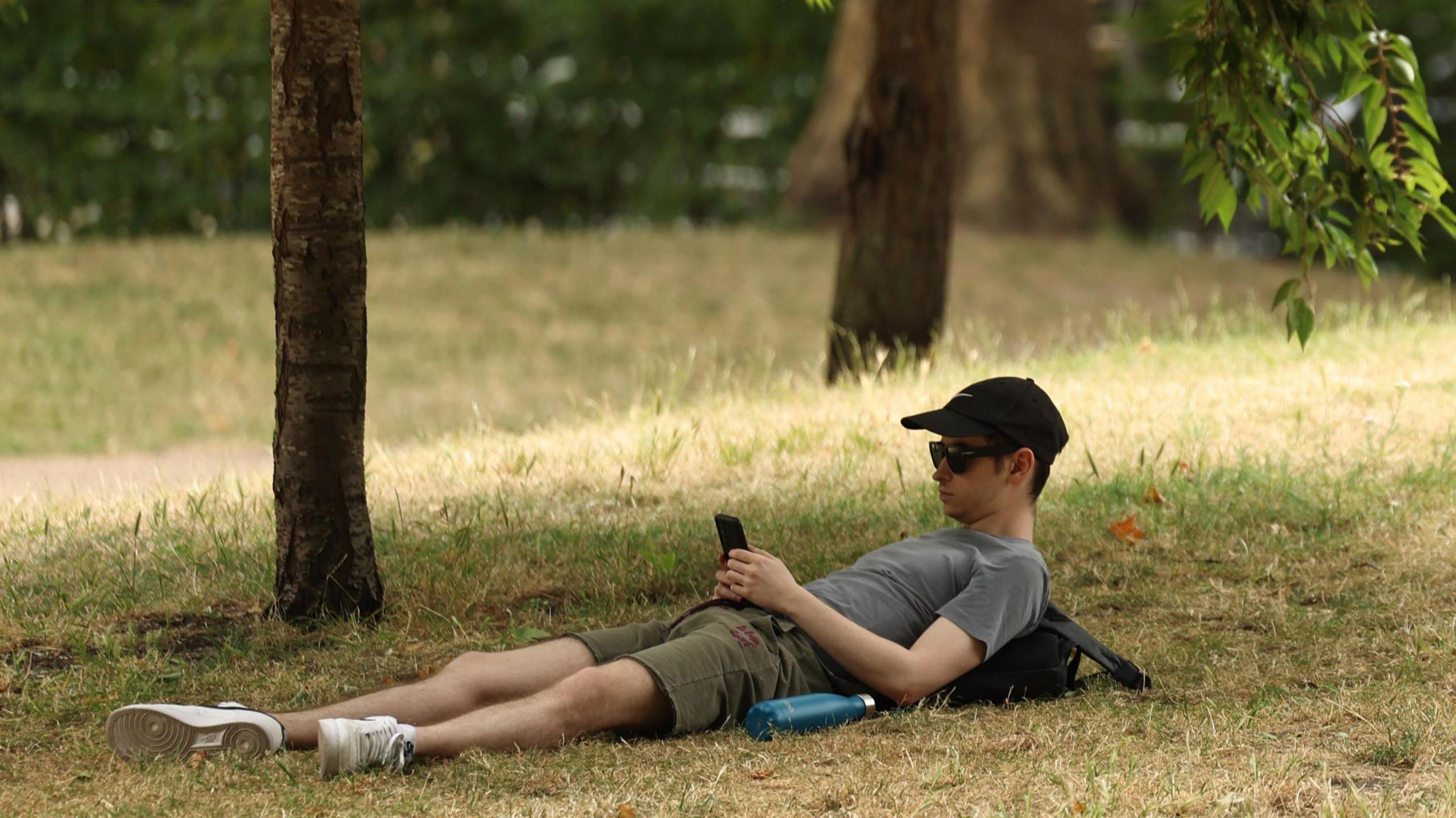 A man wearing a baseball cap and sunglasses using a mobile phone while lying down on yellow dry-looking grass, under trees