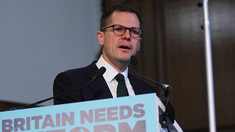 Headshot of Jenrick who speaks into two small microphones on stands on a podium in front of him that has a blue sign partially cut out of the frame, but you can read it says 'Britain needs reform'.