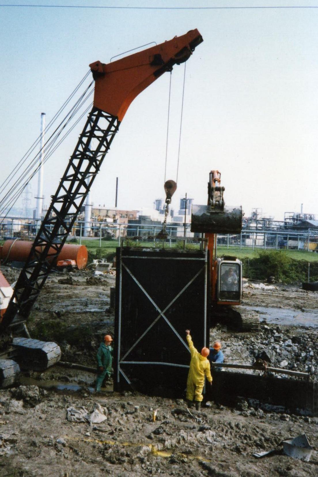 A work site where a crane is lowering something in a black container into a hole. There are workers wearing overalls and orange hats stood around the site.