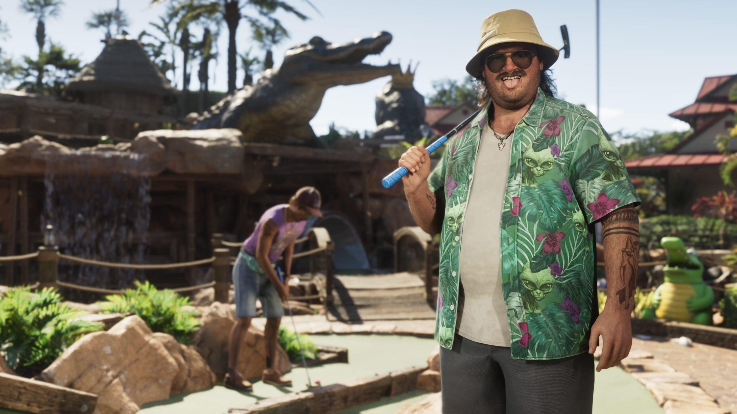 Cal Hampton, a character from GTA 6, stands in front of an alligator-themed crazy golf course. He holds a putter over one shoulder, and pulls a tongue-out face at the viewer. His loud, green, floral shirt stands out against the browns of the golf course. Behind him, a companion can be seen lining up an attempted putt, while a large alligator statue looms over the course.