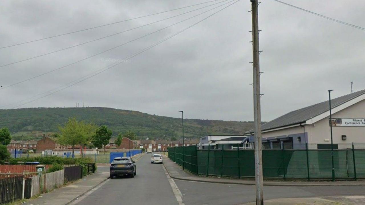 Derwentwater Road between Grangetown and Eston near Middlesbrough. On the right of the residential street is a green wired fence in front of a community hall. On the left is some gardens, then a children's park, with an estate containing two-storey homes further along. There is a rolling hill in the distance which is green with large areas of woodland.