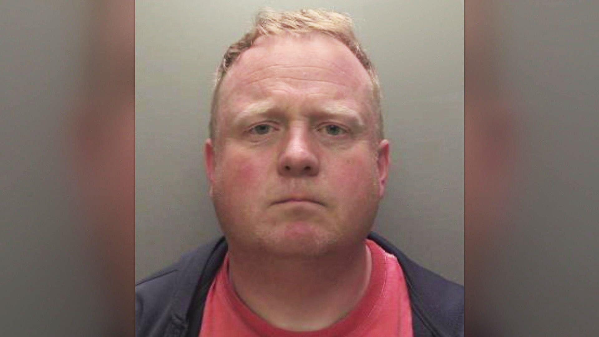 Robert Bush with short, light brown hair is shown in a straight‑on mugshot photograph. He has a neutral expression and is wearing a dark jacket over a red shirt. The background is plain and grey.