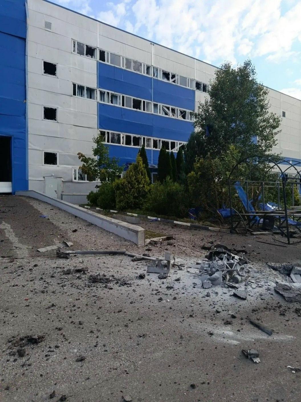 The windows of a large industrial building are smashed, with the tarmac in front charred and damaged. Debris is littered on the ground.
