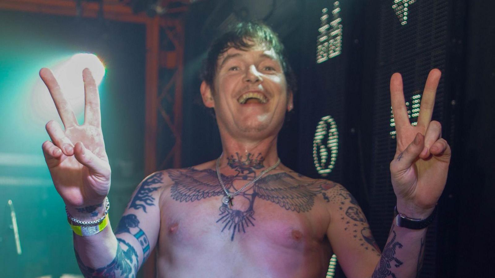 Derry Brownson of EMF smiles as he holds both hands in peace signs while shirtless at a performance