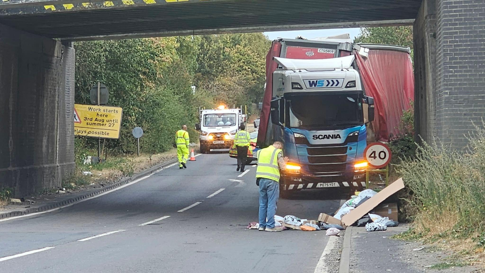 A man stands in front of a damaged lorry that has crashed into the bridge.
