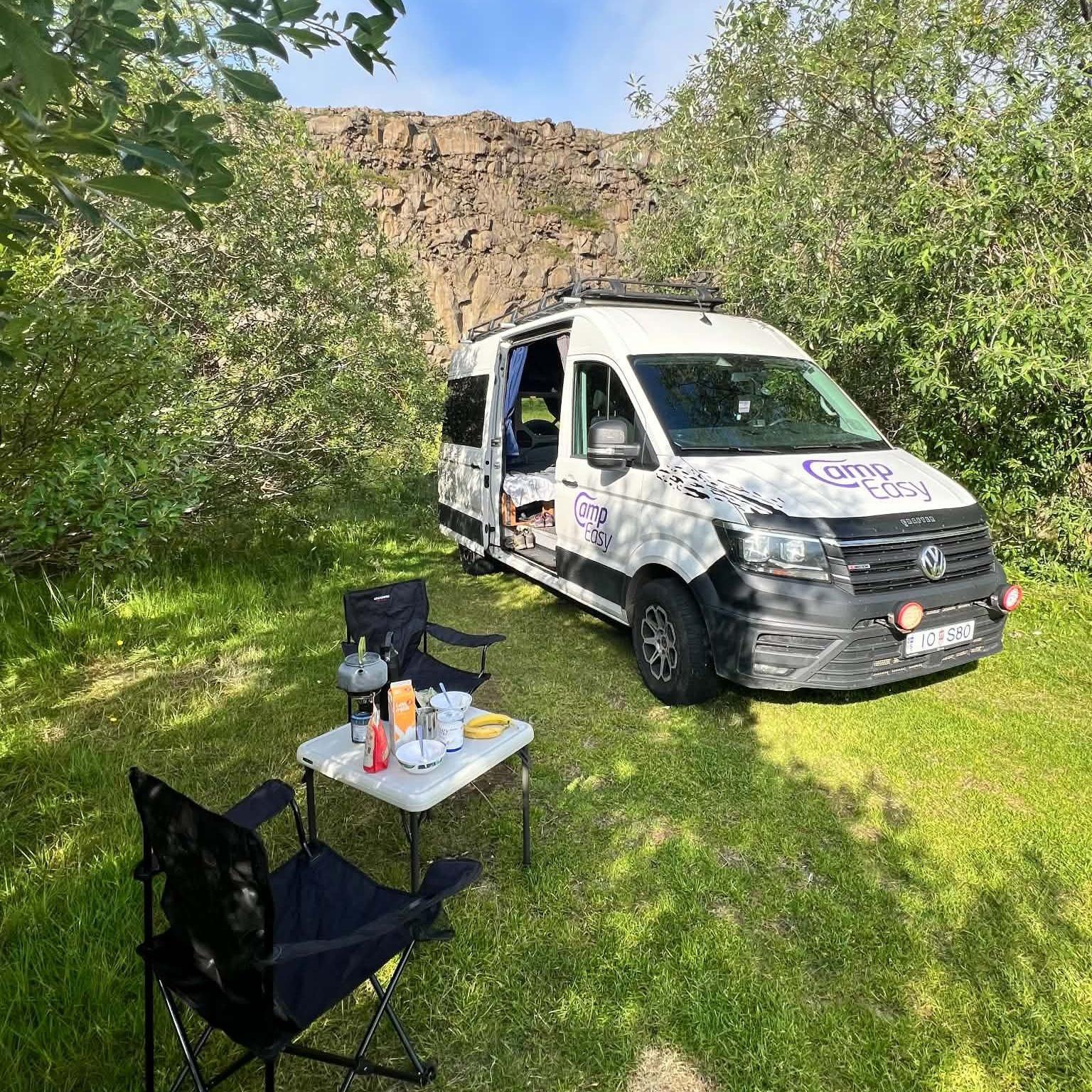 White camper van displaying parked on grass in the foreground, with the side door open. In front of the van, two folding camping chairs and a small table hold a kettle, bowls, cups and food items. Trees surround the campsite, and a rocky cliff rises behind the van. A roof rack is mounted on top of the vehicle.