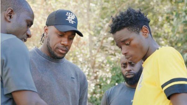 Michail Antonio, wearing a heavy grey T-shirt and a black baseball cap watches on as another man issues instruction to a footballer wearing a yellow shirt. Another man is visible in the back of the shot, with all four men looking down and fixing their gaze at something which the instructor is showing off-camera