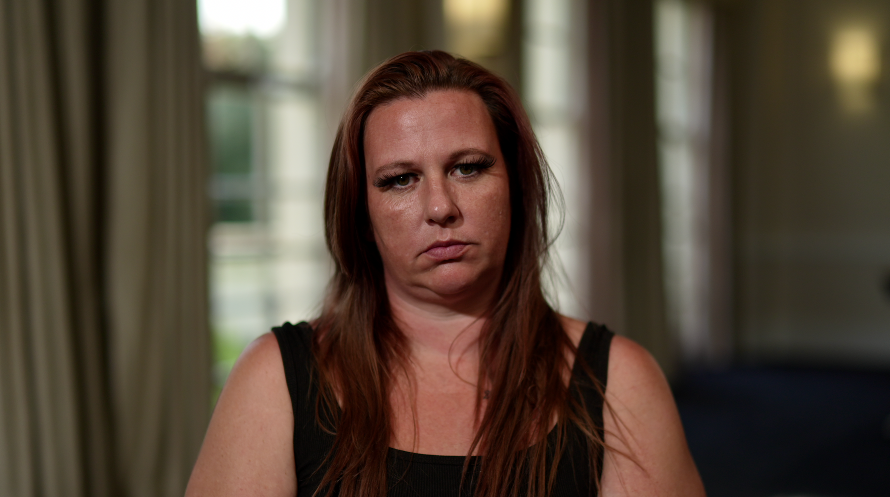 A woman wearing a black tank top and long reddish brown hair sits and stares at the camera