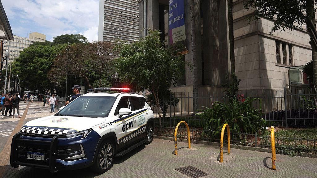 A police car with flashing lights stands in  front of the main entrance to the Biblioteca Mario de Andrade in São Paulo. A uniformed police officer is standing guard next to the entrance. A banner hanging from the column at the left-hand side of the entrance advertises the exhibition "From book to museum".
