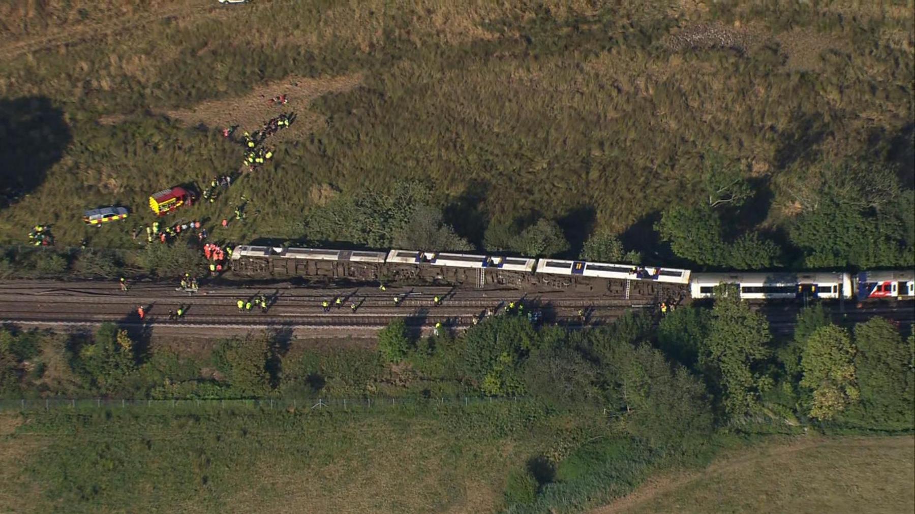 An aerial photograph shows a passenger train with multiple derailed carriages near Lewes in East Sussex. Carriages are off the tracks, with some lying on their side beside the railway. Emergency service vehicles and dozens of responders can be seen on the railway line and in a nearby field surrounded by trees and open countryside.