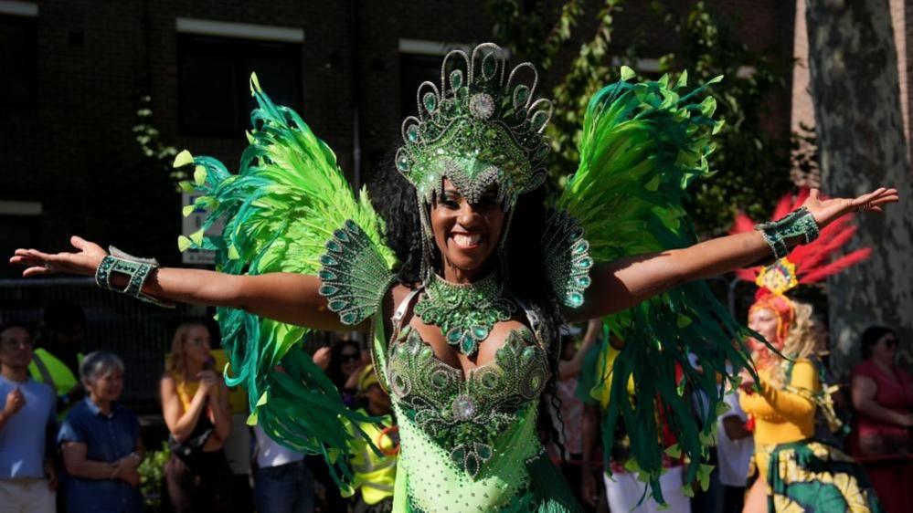 A dancer in green with a flurry of feathers behind her holds her arms out with a wide smile.